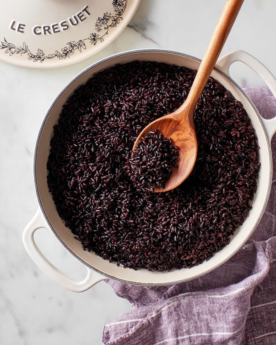 A white shallow round dish filled with a single layer of dark black rice that looks soft and dense. A wooden spoon with a smooth finish is partially resting on the rice, positioned on the right side of the dish with the handle pointing outwards. The dish is placed on a white marbled surface with a purple cloth with light stripes partially underneath it on the bottom right side. The top of a white lid with the word
