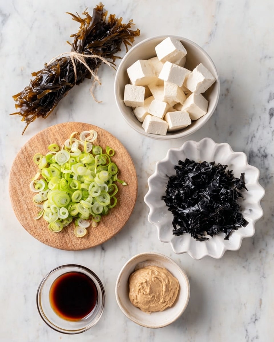 The image shows six items arranged on a white marbled surface. There is a small tied bundle of dark brown dried kelp on the top left. To its right, a white bowl holds small cubes of white tofu. Below the kelp, a small round wooden board holds a layer of thin green spring onion slices, light and dark green in color, spread evenly. Next to the tofu, on the right, is a white scalloped dish filled with black seaweed pieces that look shiny and slightly curled. Below the seaweed, a smaller white bowl contains a light brown, smooth paste. In front of this bowl, a small clear glass cup holds a dark brown liquid. Photo taken with an iphone --ar 4:5 --v 7