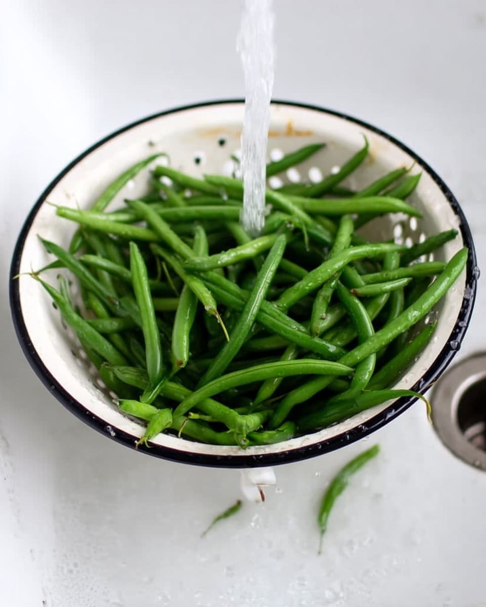 A white colander with a black rim is filled with fresh green beans being washed under running water. The beans are long and slender with a bright green color. The colander sits in a white sink, and the background is a white marbled texture. The green beans fill the colander evenly, showing their smooth texture and fresh look. photo taken with an iphone --ar 4:5 --v 7
