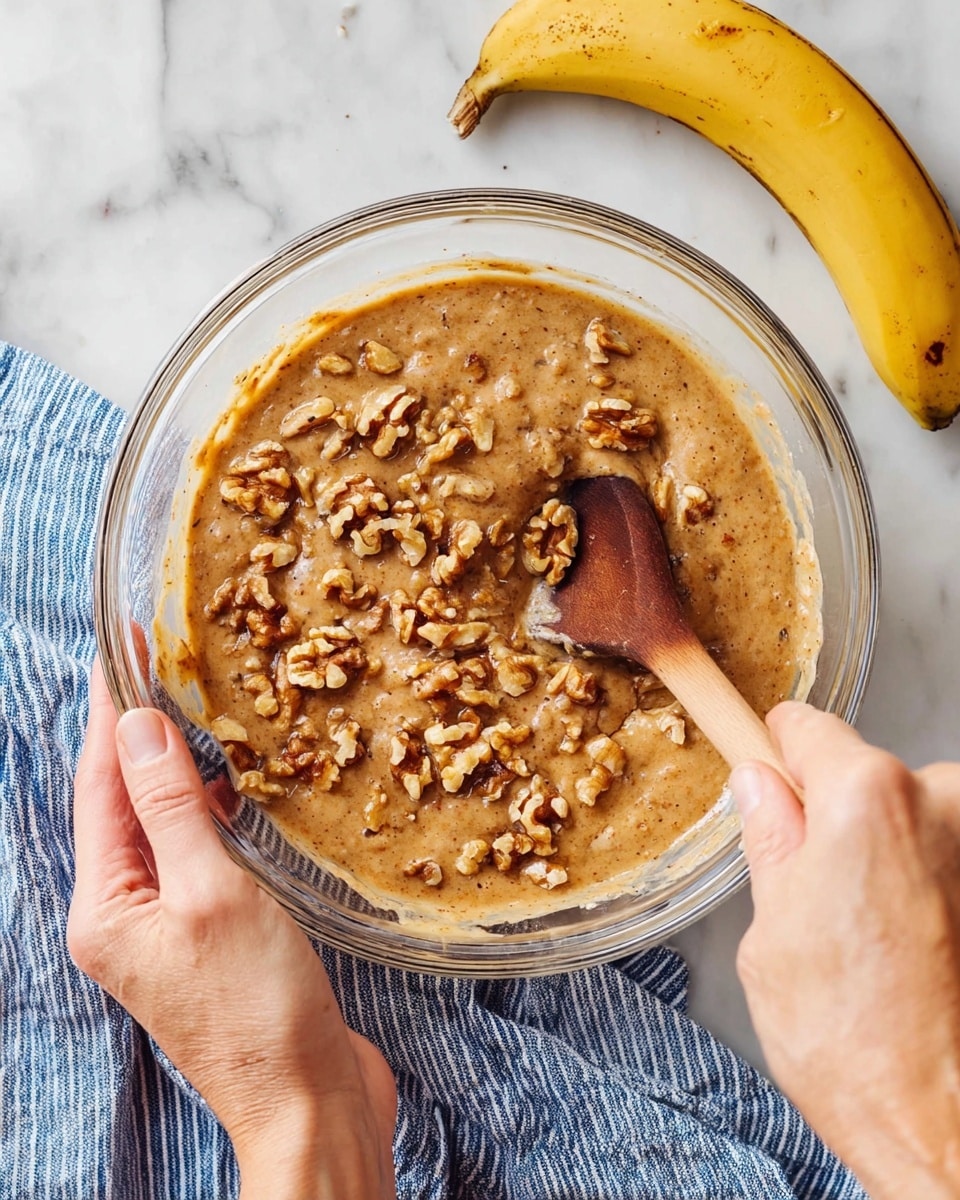 A clear glass bowl on a white marbled surface holds light brown, thick batter mixed with medium-sized walnut pieces, creating a textured look with chunks scattered evenly. A wooden spoon with a darker tip stirs the mixture, held by a pair of woman's hands, one steadying the bowl and the other mixing. A ripe yellow banana with brown spots lies near the bowl, adding color contrast. A blue and white striped cloth sits beside the bowl. Photo taken with an iphone --ar 4:5 --v 7