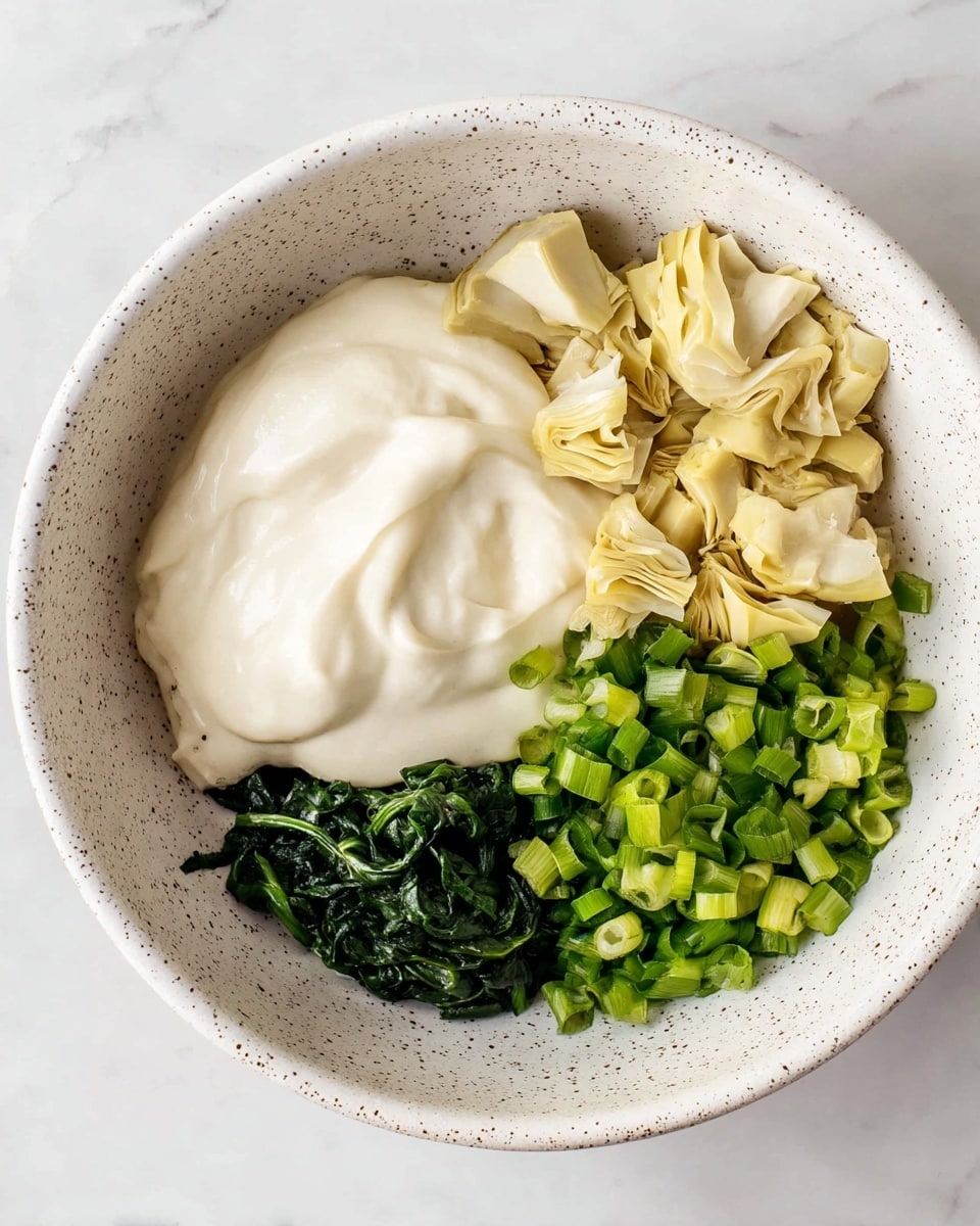 A white bowl with a speckled design holds three main layers of ingredients. On the left is a large dollop of smooth, creamy white sauce with a soft texture. On the top right, there are pale yellow pieces of chopped artichoke hearts with a slightly fibrous look. Below them, on the bottom right, is a mix of chopped bright green spring onions with a crisp texture and a small pile of dark green cooked spinach leaves. The bowl sits on a white marbled surface. photo taken with an iphone --ar 4:5 --v 7