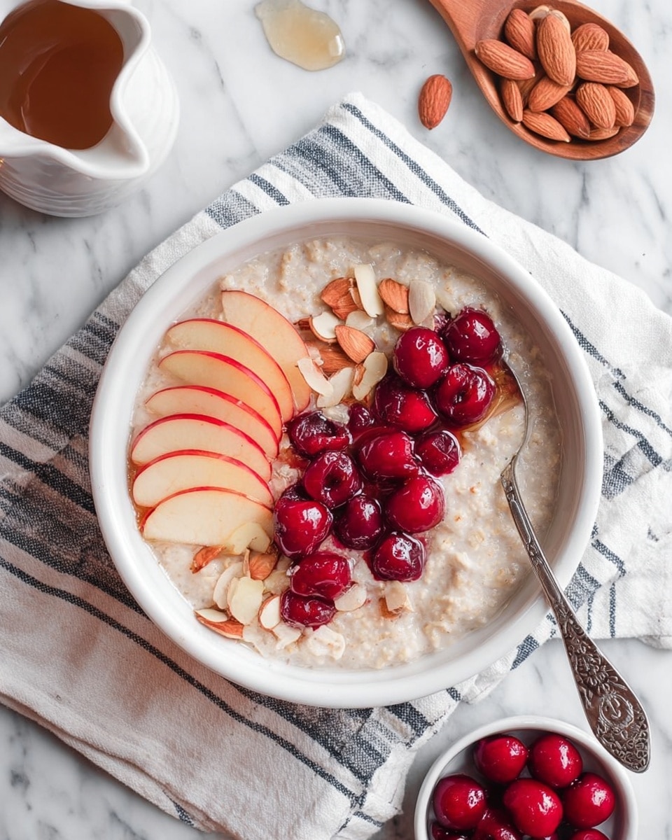A white bowl filled with creamy oatmeal as the base layer, topped with a neat row of thin, pale apple slices with red edges placed on one side. Bright red cherries in syrup form a small heap next to the apple slices, their shiny surfaces contrasting with the softness of the oatmeal. Scattered roughly around the fruit are small chunks of light brown almonds. A silver spoon with intricate designs rests inside the bowl on the right side. The bowl sits on a cloth with white and dark blue stripes, all placed on a white marbled surface. A small white jug with syrup is visible in the top left corner, along with a wooden spoon holding whole almonds and a small white bowl of red cherries near the bottom of the image. Photo taken with an iphone --ar 4:5 --v 7