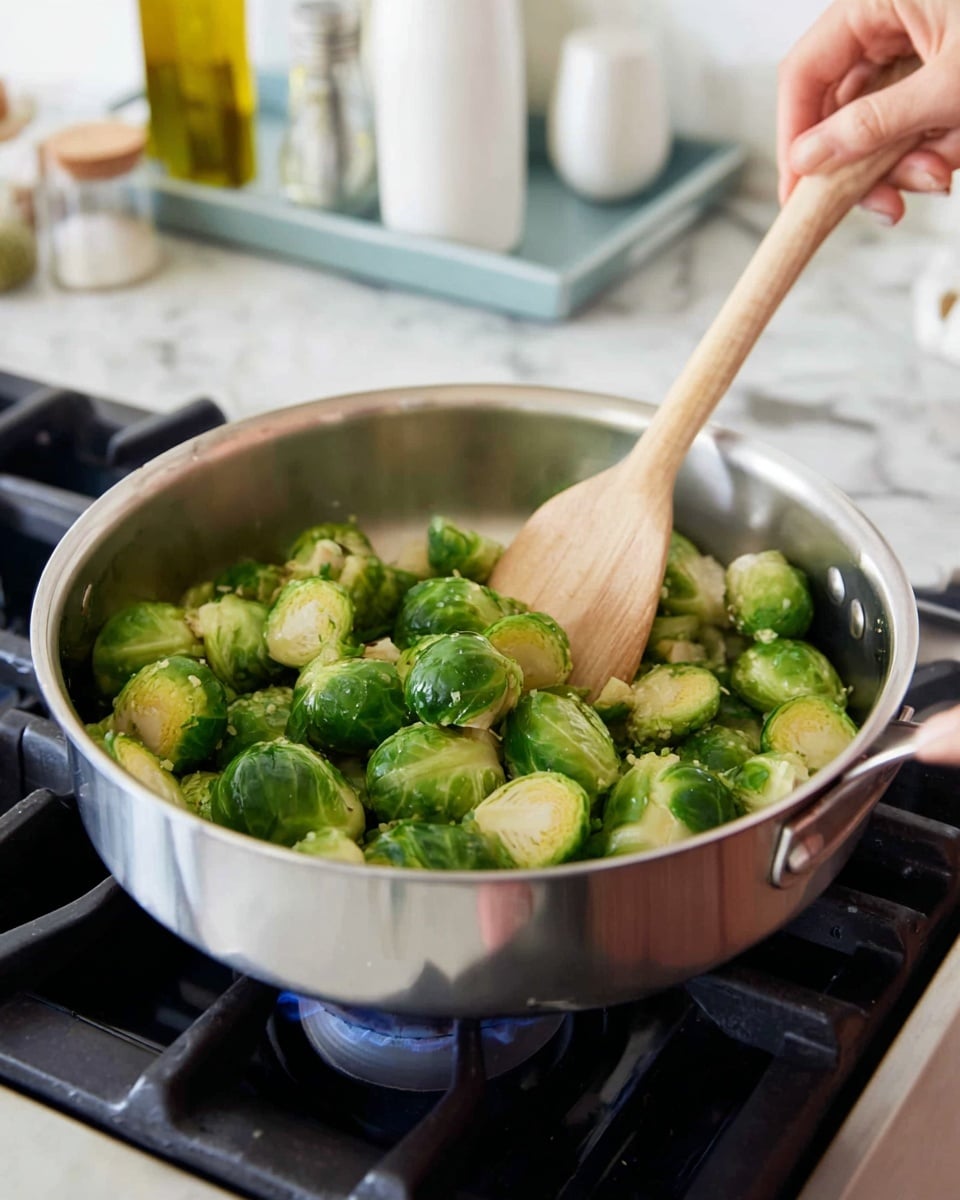 A close-up image shows a woman's hand stirring a pan of halved Brussels sprouts with a light brown wooden spoon. The Brussels sprouts are bright green with fresh, slightly glossy leaves, filling the shiny metal pan about halfway. The pan is placed on a black stove with visible blue gas flames underneath. The background includes a white marbled countertop with some kitchen items blurred out, including a light blue tray holding a small bottle of olive oil and white containers. photo taken with an iphone --ar 4:5 --v 7