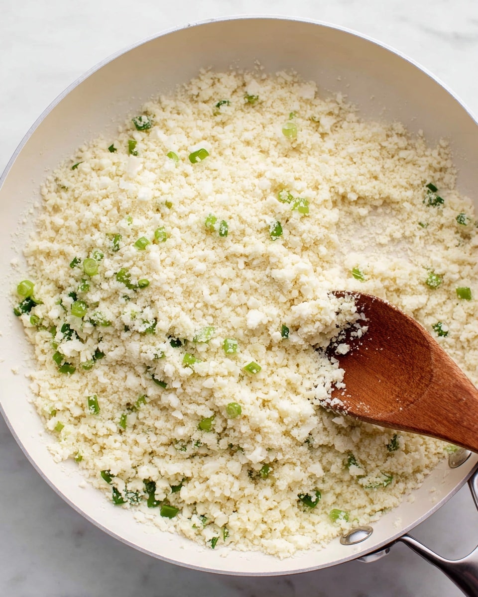 A white pan filled with finely crumbled cauliflower pieces mixed with small green onion bits. The cauliflower has a grainy, fluffy texture and is light cream in color, while the green onions add small bright green pops throughout. A wooden spoon is partly dipped into the cauliflower mixture, stirring it from the bottom right side of the pan. The pan sits on a white marbled surface. photo taken with an iphone --ar 4:5 --v 7