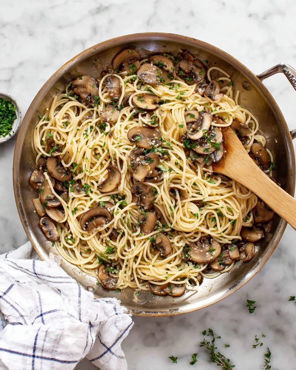 A silver pan filled with cooked spaghetti pasta mixed with sautéed brown mushroom slices and sprinkled with chopped green herbs, resting on a white marbled surface with a white cloth with blue checks near the pan. A light wooden spatula is in the pan moving through the pasta, and some scattered herbs are around the pan. Photo taken with an iphone --ar 4:5 --v 7