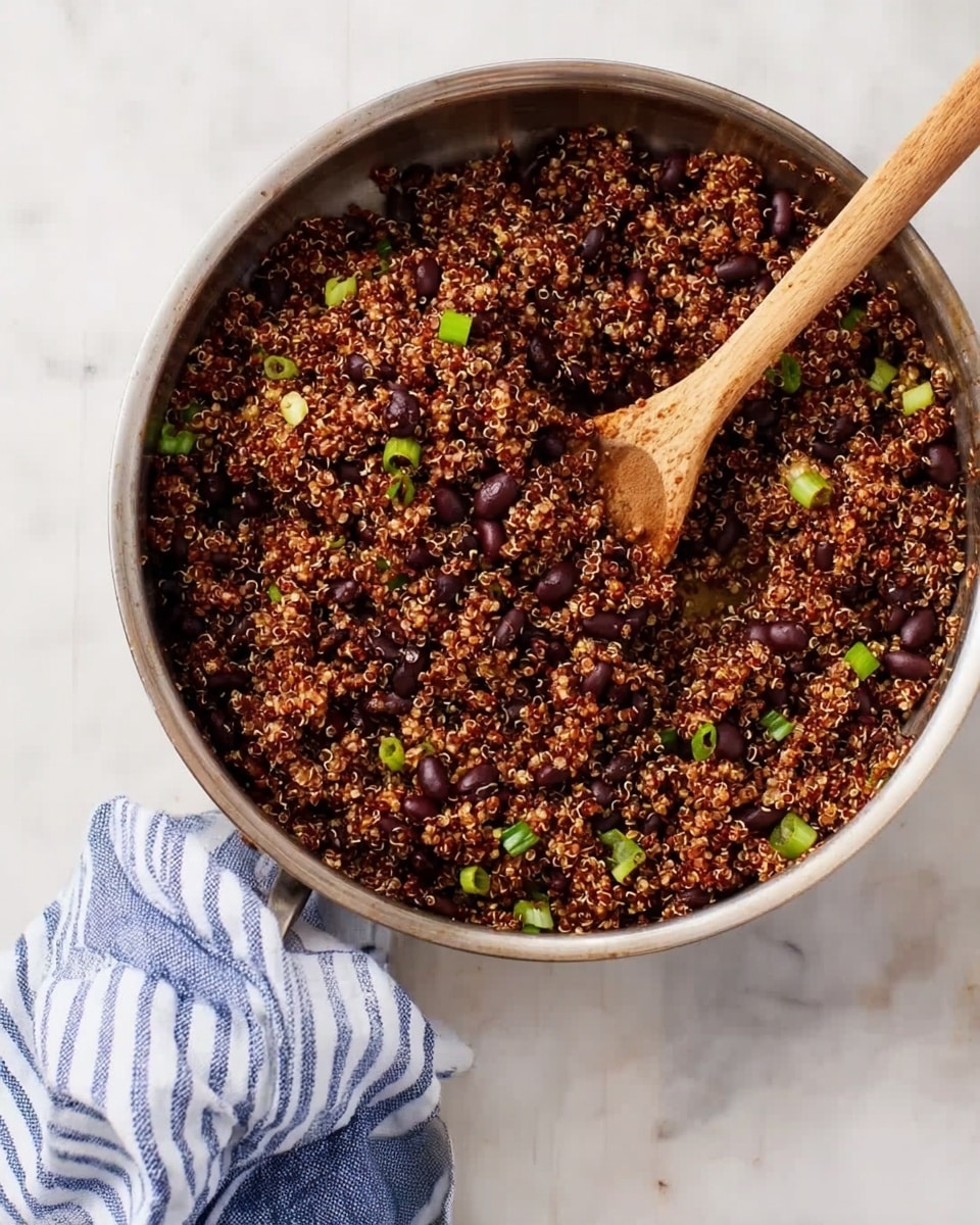 A round metal pan filled with cooked red quinoa mixed with black beans and small green onion pieces. The quinoa looks fluffy with tiny spiral grains, and the beans are scattered evenly on top. There is a wooden spoon placed inside the pan, resting on the quinoa mix. A striped blue and white cloth lies partly around the edge of the pan. The pan sits on a white marbled surface, giving a clean, simple background. photo taken with an iphone --ar 4:5 --v 7