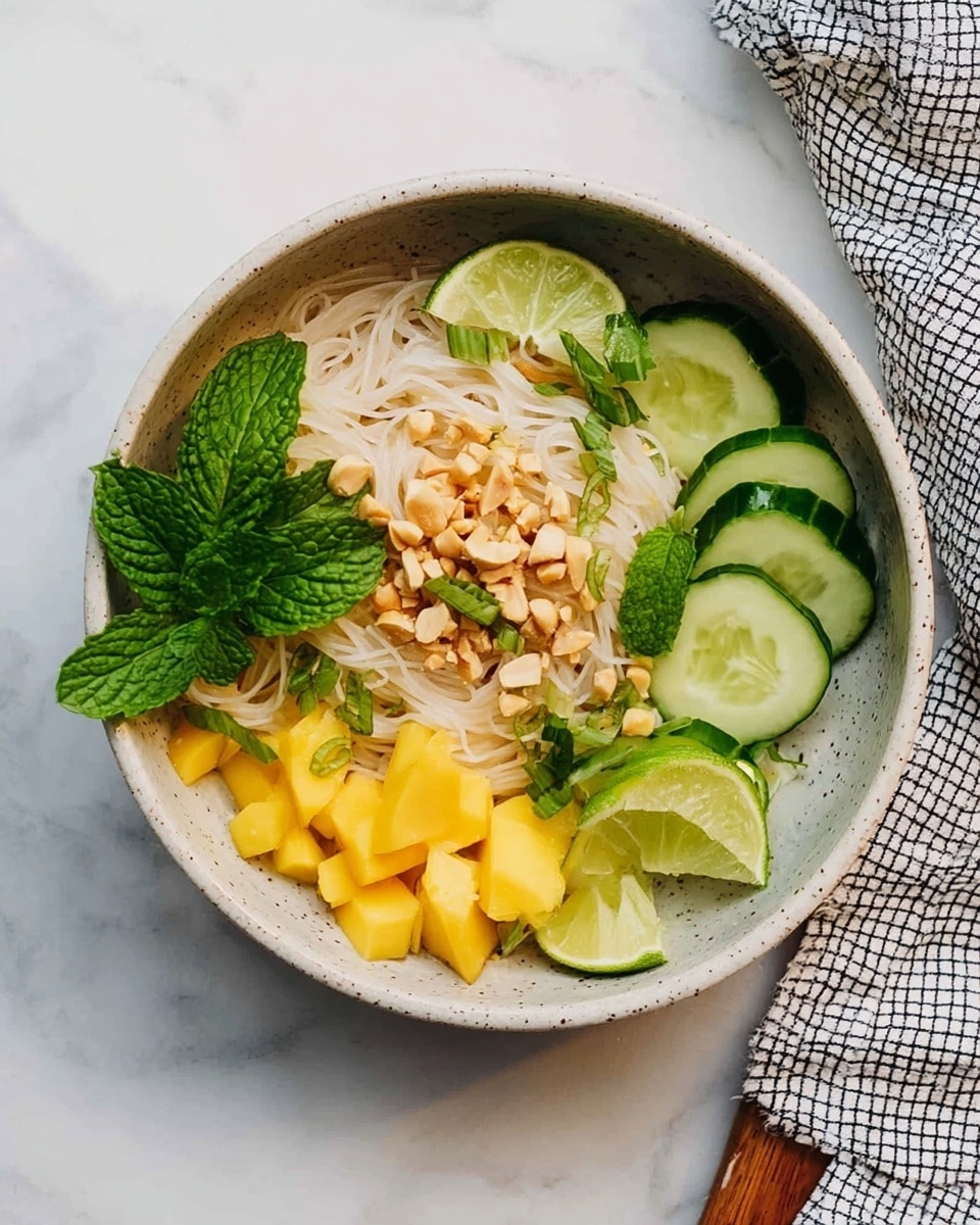 A bowl with thin white noodles at the center forms the first layer, topped with a light tan sauce and chopped nuts. Around the noodles, there are bright green cucumber slices arranged at the right side and small yellow mango cubes at the bottom left. Two fresh mint leaves sit on top of the noodles. A lime wedge is placed near the cucumbers. The bowl is light gray with speckles and is set on a white marbled surface with part of a checkered cloth on the right side. Photo taken with an iphone --ar 4:5 --v 7