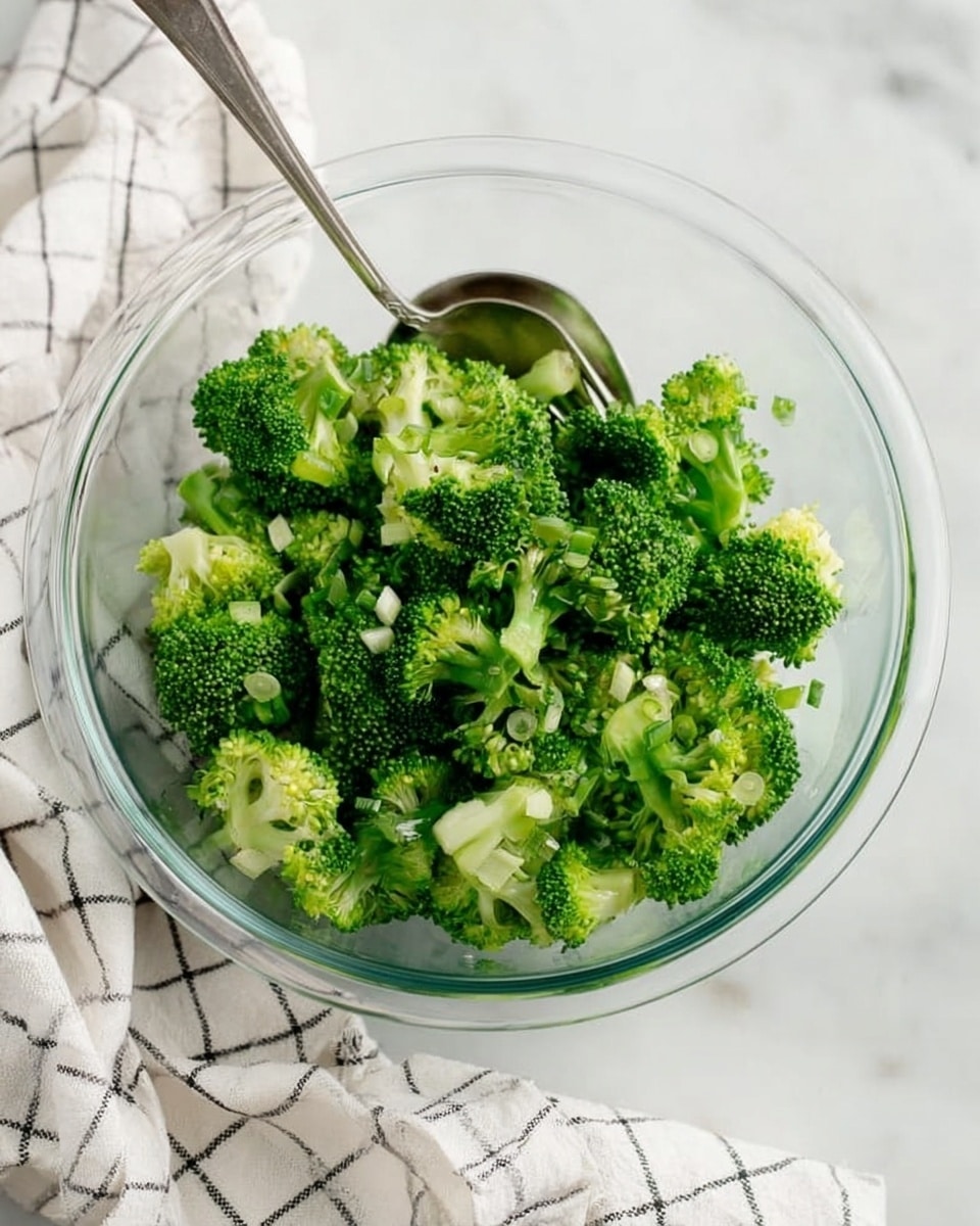 A clear glass bowl filled with small, bright green broccoli florets that have a fresh, rough texture. The broccoli pieces are uneven in size, layered in one group inside the bowl. There are tiny chopped green onions mixed in with the broccoli, adding small dots of lighter green. A silver spoon sits inside the bowl, partly under the florets with its handle pointed to the upper left. The bowl is placed on a white marbled surface, next to a white cloth with a black checked pattern. The cloth is casually folded and rests to the right and below the bowl. Photo taken with an iphone --ar 4:5 --v 7