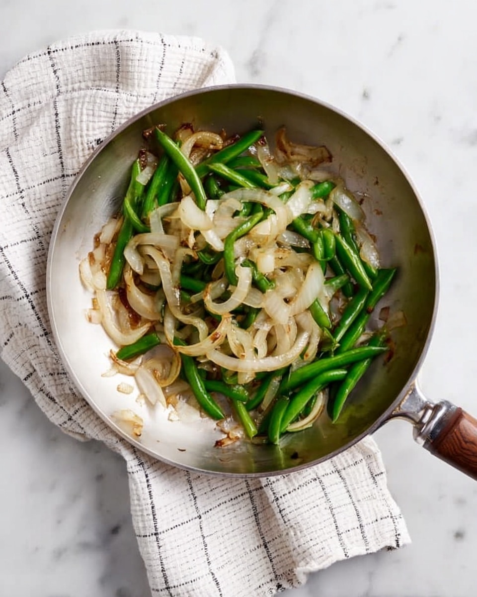 A silver frying pan is placed on a white marbled surface with a white and black checkered cloth under its handle. Inside the pan, there are cooked light brown and slightly soft sliced onions mixed with bright green cooked snap peas. The vegetables are spread unevenly across the pan, showing some light browning on the onions. The background is clean and simple, focusing on the pan and its contents. photo taken with an iphone --ar 4:5 --v 7