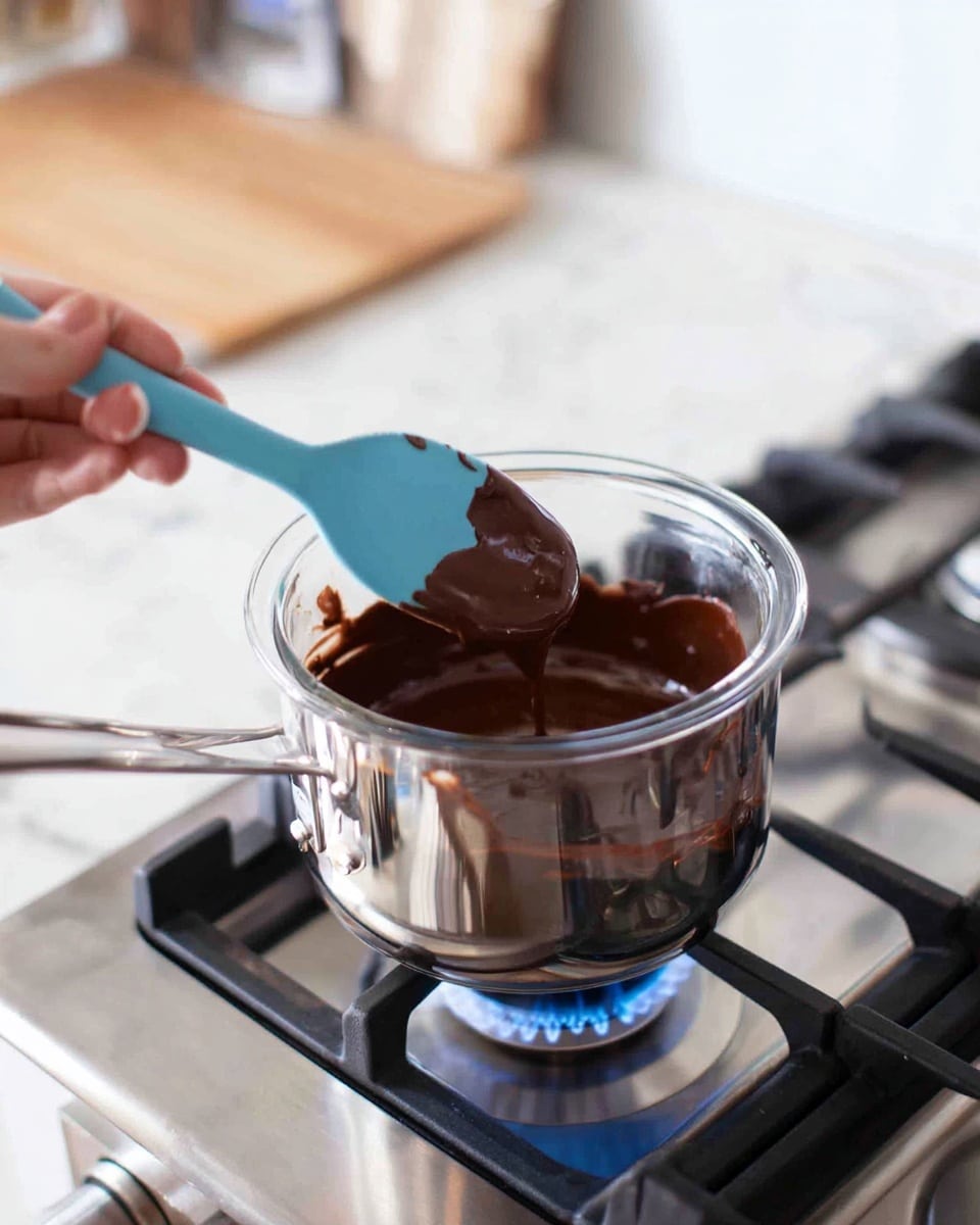 A clear glass bowl filled with thick, dark brown melted chocolate sits in a shiny metal pot on a gas stove blue flame. A woman's hand is holding a blue spatula, stirring the smooth chocolate with a glossy texture inside the bowl. The kitchen surface around the stove is white with a marbled texture, and blurred objects like a white phone and a wooden cutting board can be seen in the background. photo taken with an iphone --ar 4:5 --v 7
