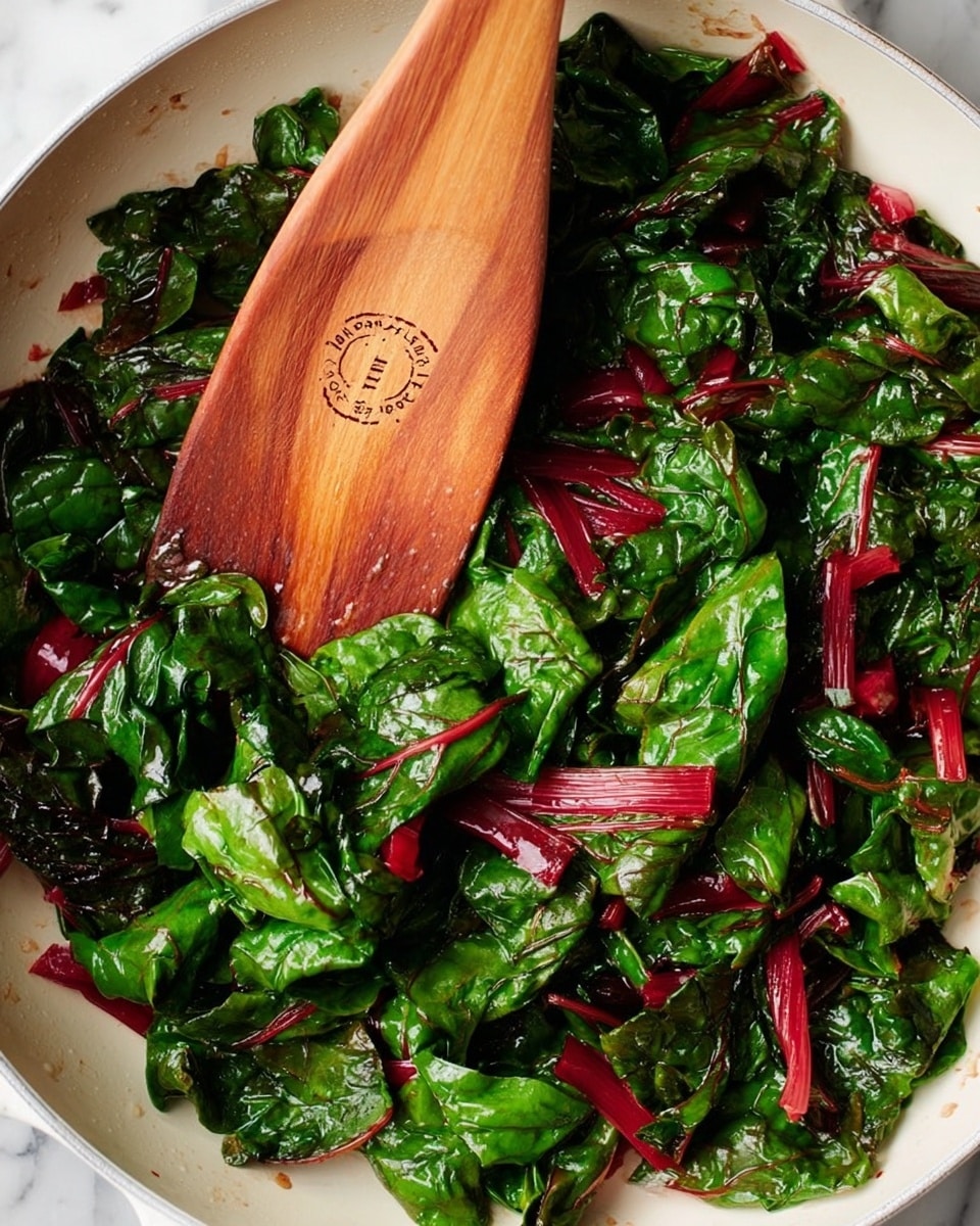 A white pan filled with bright green and deep red leafy greens that have a shiny, oily texture, suggesting they are cooked or sautéed. The greens have thick red stems and crinkled leaves layered unevenly across the pan. On top of the greens lies a smooth, light brown wooden spatula with an engraved circle and text. The background is a white marbled surface. Photo taken with an iphone --ar 4:5 --v 7
